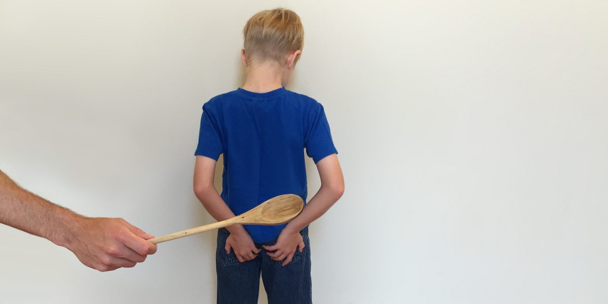 A boy stands facing a wall, hands on his buttocks, while a wooden spoon is held up behind him as if to spank.