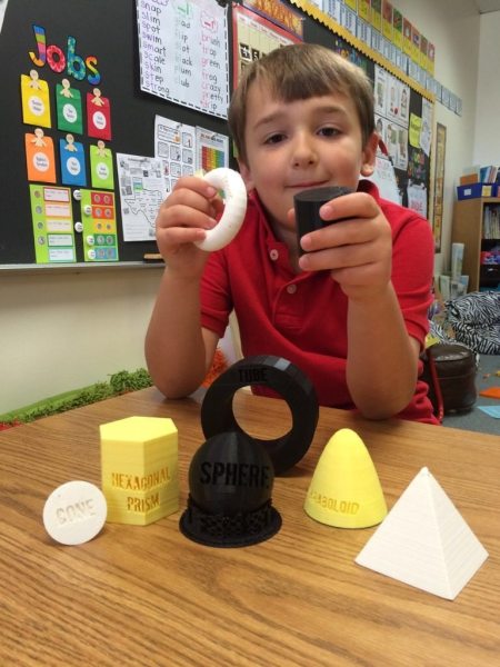 1st grader holds two 3D shapes while others sit on a table in front of him