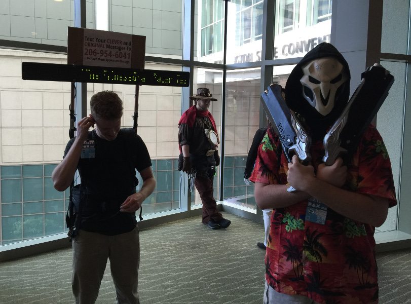 Overwatch cosplay from PAX West 2016. A reaper in a Hawaiian shirt is close to the camera at the front right, guns across his chest, with a McCree leaning up against a glass wall in the background. To the left is a non-cosplay person with a LED display mounted on his back advertising for people to send him their original text message, which will be displayed on the LED board.