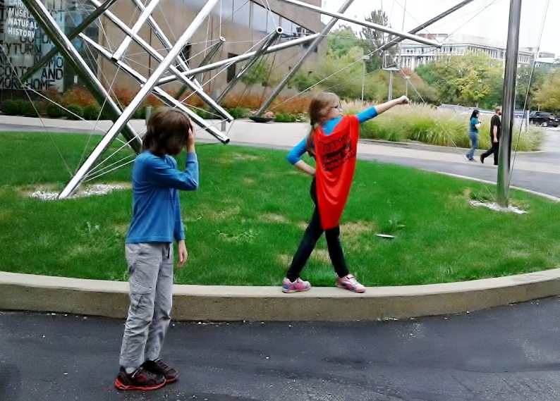 Girl in "Reading Gives You Superpowers" cape poses in front of an outdoor sculpture, her brother looks embarrassed.