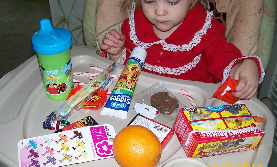 toddler with high chair tray full of stocking stuffers including chocolate, toothpaste, a toothbrush, barrettes, an orange, and animal crackers