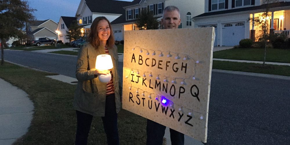 The author holding a board with fabric wallpaper and lights, with a letter painted beside each light from A to Z. A woman dressed as Joyce from Stranger Things is to the left.