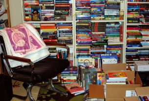 Photo of a chair with a blanket draped over it, next to three overstuffed bookshelves, along with piles of books and boxes of books on the floor next to it.