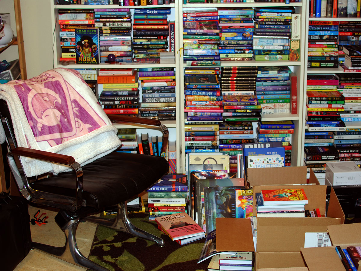 Photo of a chair with a blanket draped over it, next to three overstuffed bookshelves, along with piles of books and boxes of books on the floor next to it.